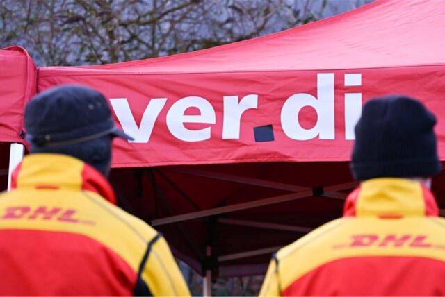 Zwei Mitarbeiter des Logistikkonzerns DHL stehen an einem Warnstreik-Posten der Gewerkschaft Verdi, der sich am Eingang einer DHL-Zustellbasis befindet.