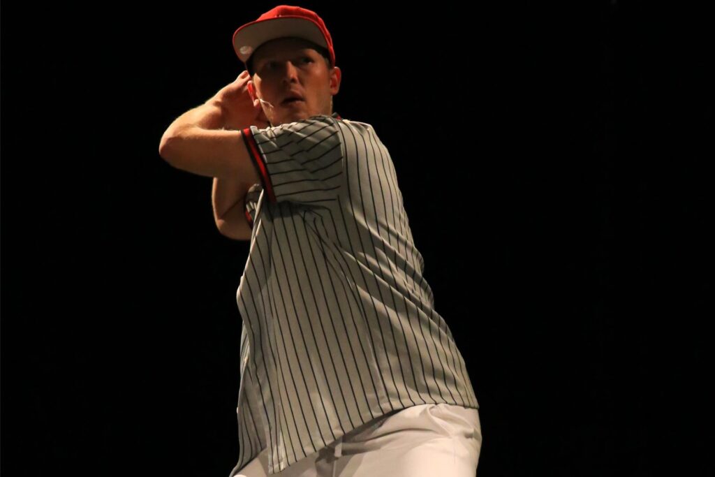 er Darsteller von „John & Jen“ in einem Baseball-Outfit auf der Musicalbühne. Das Baseball-Shirt ist grau mit schwarzen Streifen und die Cap ist rot und die Hose weiß.