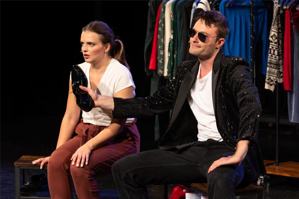 Die beiden Darsteller von „John & Jen“ werden alleine auf der Bühne der Rausinger Halle stehen. Beide tragen ein weißes Shirt. Sie trägt eine rote Hose und er eine schwarze Hose sowie ein schwarzes Sakko. Er hat einen Schuh in der Hand und trägt eine schwarze Sonnenbrille.