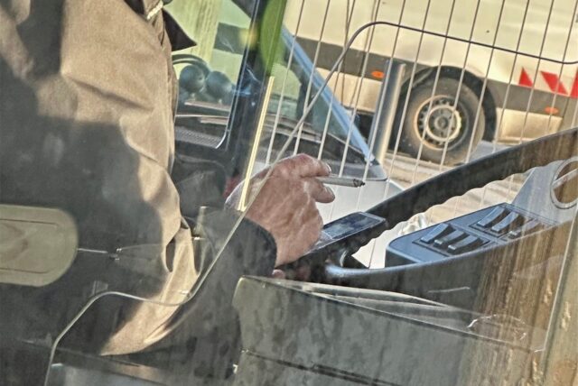 Das Lenkrad eines Busses ist zu sehen, davor die Hand des Fahrers mit einer Zigarette darin.