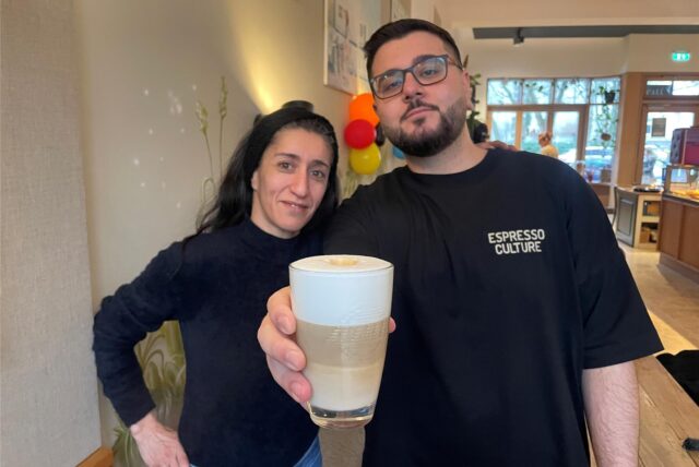 Moe und Ferda lächeln in die Kamera. Moe hält einen Kaffee in der Hand.
