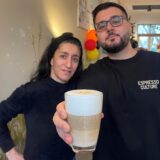 Moe und Ferda lächeln in die Kamera. Moe hält einen Kaffee in der Hand.