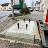 Aus einem Betonfundament in einem Gehweg in Unna ragen drei schwarze Metallstangen. Daneben steht eine Bake