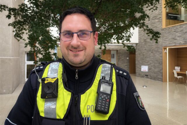 Polizeihauptkommissar Dennis Kischel ist der neue Bezirksdienstbeamte in Holzwickede. Hier steht er im Atrium des Holzwickeder Rat- und Bürgerhauses. Er trägt eine Polizei Uniform und eine Brille.