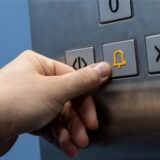 Eine Hand drückt im Fahrstuhl auf einen Notfallknopf mit einer gelben Glocke.