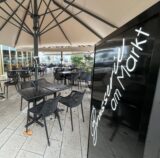 Die Terrasse vor dem Eingang der Brasserie am Markt in Holzwickede. Ein großer beiger Schirm mit mehreren schwarzen Tischen und Stühlen. Rechts befindet sich das schwarz-weiße Eingangsschild „Brasserie am Markt..