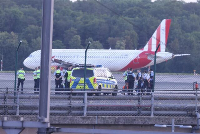 Sicherheitspersonal und Polizisten stehen auf dem Flugfeld und versuchen Aktivisten der Gruppe Letzte Generation vom Asphalt zu lösen, nachdem sie sich festgeklebt haben.