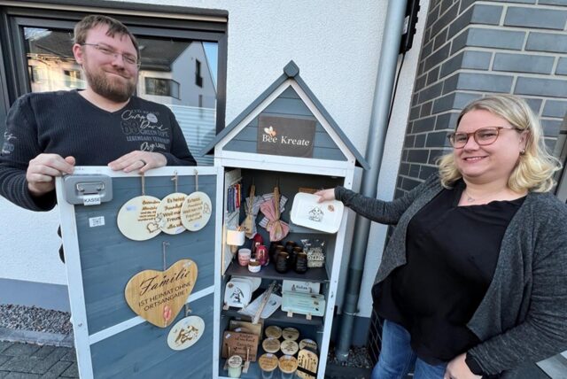 Philipp Bien (links) und Katja Lösch bieten in ihrem Bee-Kreate-Schrank seit Februar handgefertigte Geschenke und Mitbringsel an. Auf dem Bild stehen sie am Schrank. Katja Lösch hält einen Teller in der Hand.