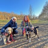 Zwei Frauen mit je einem Hund Hocken auf einer Freifläche in Unna. Im Hintergrund stehen Bäume