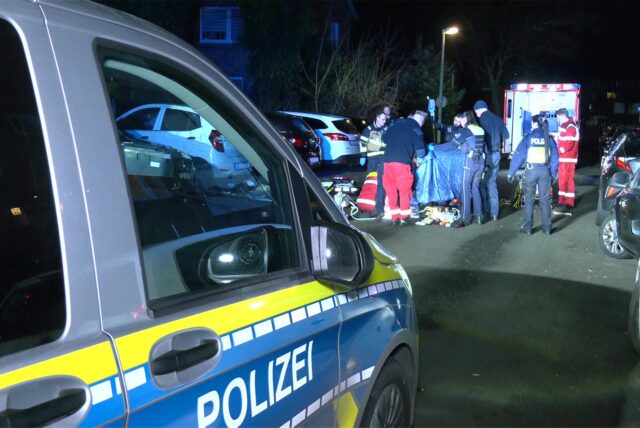 Rettungskräfte und Polizisten schirmen den Tatort mit einem Tuch ab. Im Vordergrund steht ein Polizeiwagen.