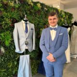 Daniel Seeliger (25) trägt einen hellblauen Hochzeitsanzug und steht neben einem ausgestellten Anzug vor einer grünen Pflanzenwand.