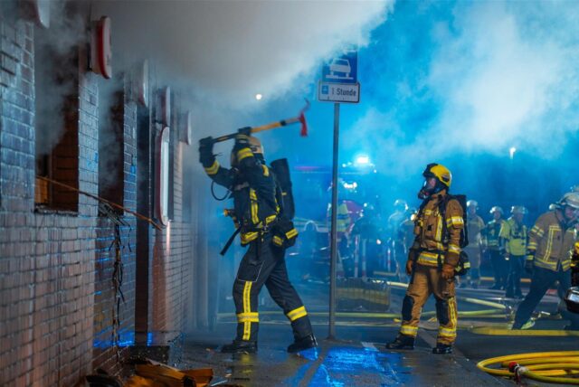 Ein Feuerwehrmann schlägt mit einer Axt die Tür einer Gaststätte in Menden auf, aus der Rauch kommt.