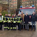 Feuerwehrleute sowie Mitarbeiter von Stadtverwaltung und Steag stehen auf dem Platz der Partnerstädte vor einem Feuerwehrfahrzeug, das vor dem Rathaus parkt.