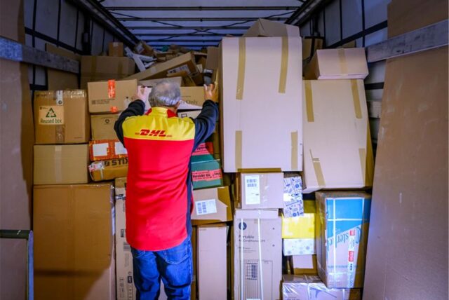 Ein DHL-Mitarbeiter steht vor einem Haufen von Paketen.