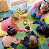 Vier Kinder sitzen mit einer Betreuerin im Kreis. Sie bestaunen eine Pflanze. Das Foto wurde aus der Vogelperspektive gemacht.