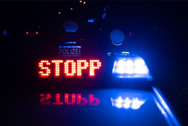 Auf dem Dach eines Streifenwagens leuchten das Blaulicht und das Schild „Stopp“. Vor dem Wagen stehen zwei Polizeibeamte.
