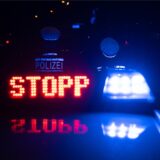 Auf dem Dach eines Streifenwagens leuchten das Blaulicht und das Schild „Stopp“. Vor dem Wagen stehen zwei Polizeibeamte.