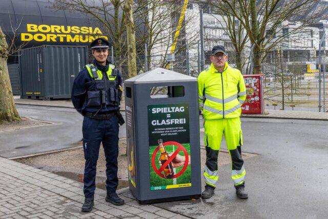 Ein Mitarbeiter vom Ordnungsamt und ein Mitarbeiter von der EDG stehen am Signal-Iduna-Park in Dortmund neben einem Glasmülleimer.