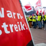 Ein Plakat mit der Aufschrift „Warnstreik“. Im Hintergrund sind Streikende zu sehen.