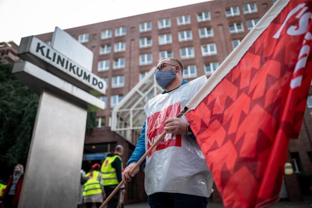 Ein Mann steht mit einer Fahne von Verdi vor dem Eingang des Klinikums Dortmund. Er trägt eine Plastikweste der Gewerkschaft Verdi, die zum Streik aufgerufen hat.