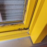 An einem postgelben Fensterrahmen sind Schäden zu sehen, die vom Versuch stammen, dieses Fenster aufzuhebeln.