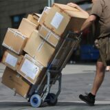 Ein Paketbote in brauner Arbeitskleidung schiebt vor sich einen Sackkarren mit einem Dutzend Paketen darauf.
