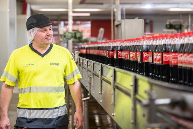 Ein Mitarbeiter von Coca-Cola steht vor einem Fließband mit Coca-Cola-Flaschen.