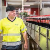Ein Mitarbeiter von Coca-Cola steht vor einem Fließband mit Coca-Cola-Flaschen.