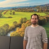 Florian Hübner, Umwelt- und Klimaschutzbeauftragter bei der Stadt Schwerte, steht vor einem Bild, das die Ruhrwiesen zeigt.