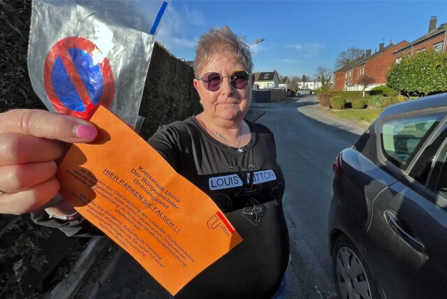 Neben einem Auto, das auf der Straße vor ihrer Tür parkt, zeigt Anwohnerin Iris Bolte einen auf orangefarbenem Papier gedruckten Warnhinweis der Stadt Unna. Autofahrer werden darauf hingewiesen, dass sie künftig mit Knöllchen zu rechnen hätten.