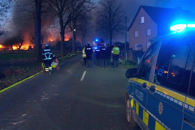 Einsatzkräfte der Polizei stehen auf der Straße vor einem brennenden Gebäude. Im Vordergrund steht ein Polizeiauto mit Blaulicht.