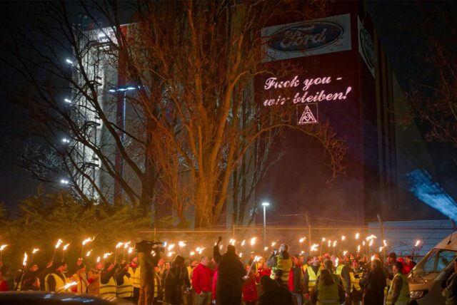 Ford-Mitarbeiter stehen mit Fackeln auf dem Gehweg vor einem Gebäude der Fordwerke, auf dem die Worte „Fuck you – wir bleiben“ projiziert wird.
