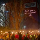 Ford-Mitarbeiter stehen mit Fackeln auf dem Gehweg vor einem Gebäude der Fordwerke, auf dem die Worte „Fuck you – wir bleiben“ projiziert wird.