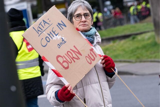 Bei einer Demonstration hält eine Person ein Schild mit der Aufschrift: Kita: Come in and burn out.