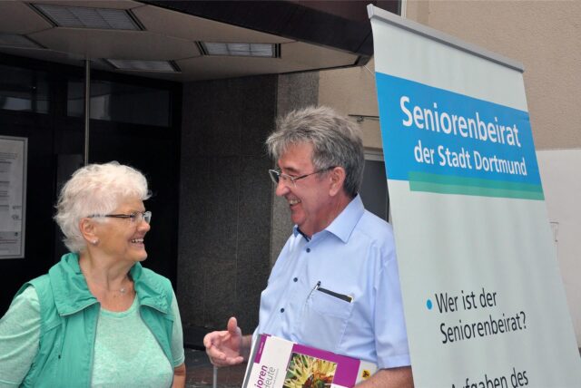 Ein Seniorenbeirats-Vertreter im Gespräch mit einer Seniorin neben einem Transparent des Seniorenbeirats.