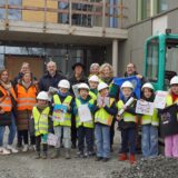 Kinder aus Unna stehen mit Baustellenhelm und Signalweste auf der Baustelle des neuen Bildungszentrums am Hertinger Tor, wo sie eine Zeitkapsel befüllt haben.