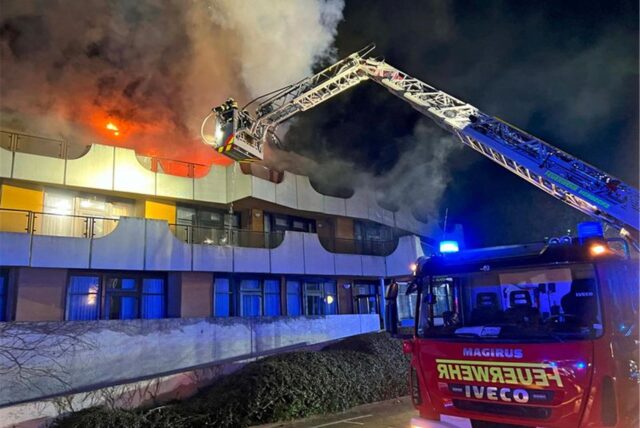 Ein Gebäude steht in Flammen – von einem Feuerwehrauto ragt ein Kran an die Flammen heran für Löscharbeiten.