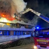 Ein Gebäude steht in Flammen – von einem Feuerwehrauto ragt ein Kran an die Flammen heran für Löscharbeiten.