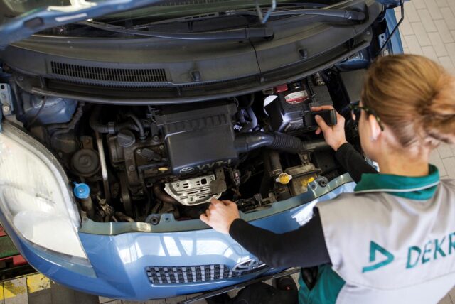 Eine Prüferin der Dekra beugt sich über den geöffneten Motorraum eines blauen Toyota Yaris.