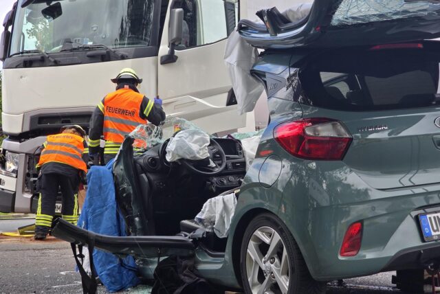 Im Vordergrund ein schwer beschädigter Kleinwagen, im Hintergrund ist ein weißer Lkw sowie Mitarbeiter der Feuerwehr zu erkennen.