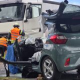 Im Vordergrund ein schwer beschädigter Kleinwagen, im Hintergrund ist ein weißer Lkw sowie Mitarbeiter der Feuerwehr zu erkennen.