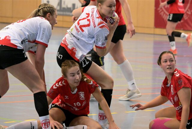 Nele Wagener vom HC Tura Bergkamen fängt einen Ball ab. Carla Verhasselt und Thia-Carolin Schmidt vom Lüner SV Handball sitzen auf dem Boden.
