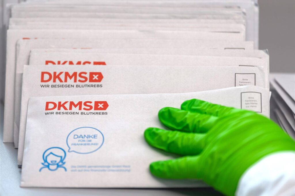 Eine Hand mit einem grünen Handschuh greift nach Briefumschlägen mit dem Aufdruck „DKMS“. Eine Hand mit einem grünen Handschuh greift nach Briefumschlägen mit dem Aufdruck „DKMS“.