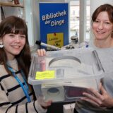 Bücherei-Mitarbeiterin Maren Austel überreicht Reporterin Janka Hardenacke eine Kunststofbox, in der sich eine Popcornmaschine befindet.