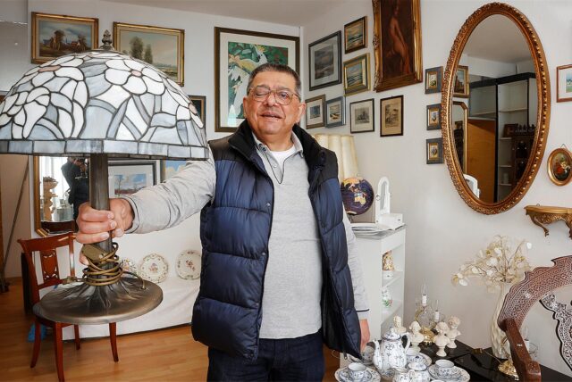 Dragan Djuric steht in seinem Antikladen und hält eine Lampe in der Hand. Im Hintergrund lassen sich Gemälde, Spiegel und Porzellan finden.