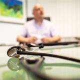 Armin Wunder, Facharzt für Allgemeinmedizin, sitzt hinter einem Stethoskop an seinem Schreibtisch in einem Behandlungszimmer seiner Praxis.