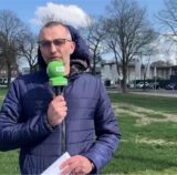 Ein Mann steht mit einem Mikrofon in der Hand auf einer Grünfläche. Im Hintergrund sind ein Gebäude, geparkte Autos und Bäume zu sehen.