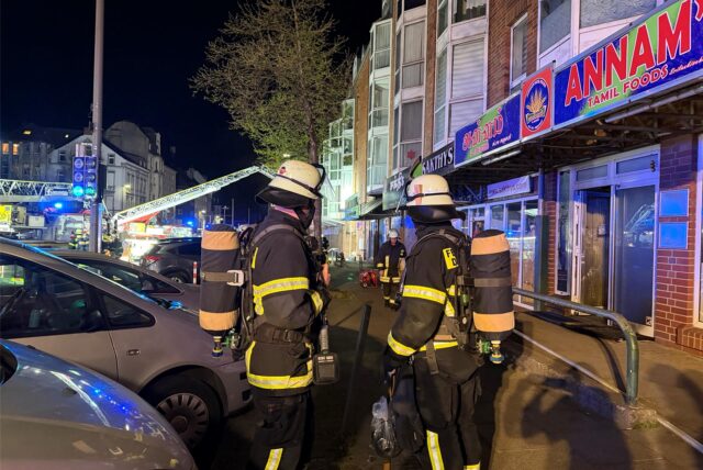 Zwei Feuerwehrmänner stehen während des Brandeinsatzes an der Rheinischen Straße in Dorztmund auf dem Bürgersteig.