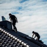 Zwei Bauarbeiter installieren eine Photovoltaik-Anlage auf einem Dach, im Hintergrund ist der bewölkte Himmel zu sehen.