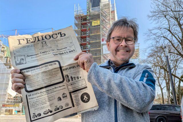 Ein Pfarrer steht vor einer eingerüsteten Kirche und hält eine alte Zeitung in den Händen.